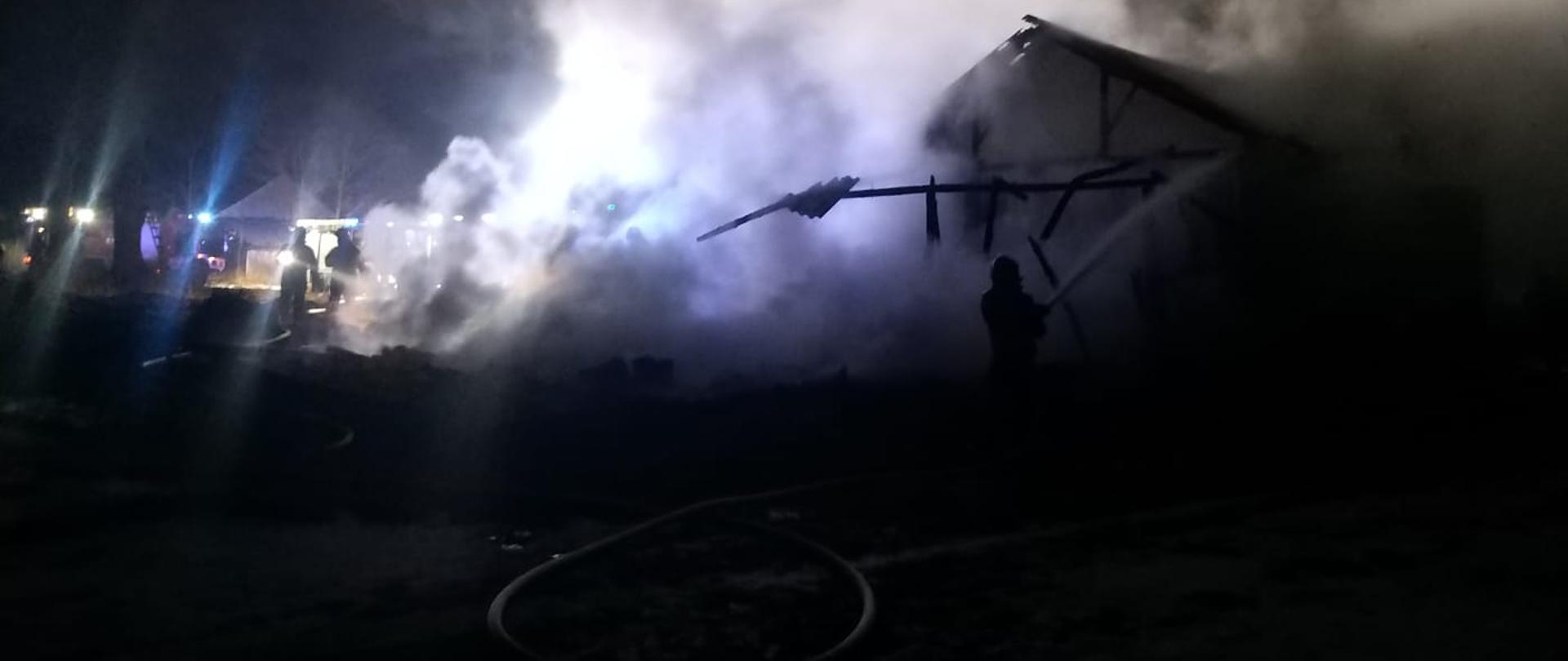 Na zdjęciu pożar drewnianej stodoły, w wyniku czego widoczny jest dym. Znajdują się również strażacy. 
