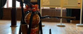 Na pierwszym planie stara pożarnicza pompa, za nią dwie tablice z opisem historycznym, w tle okna i zasłony.