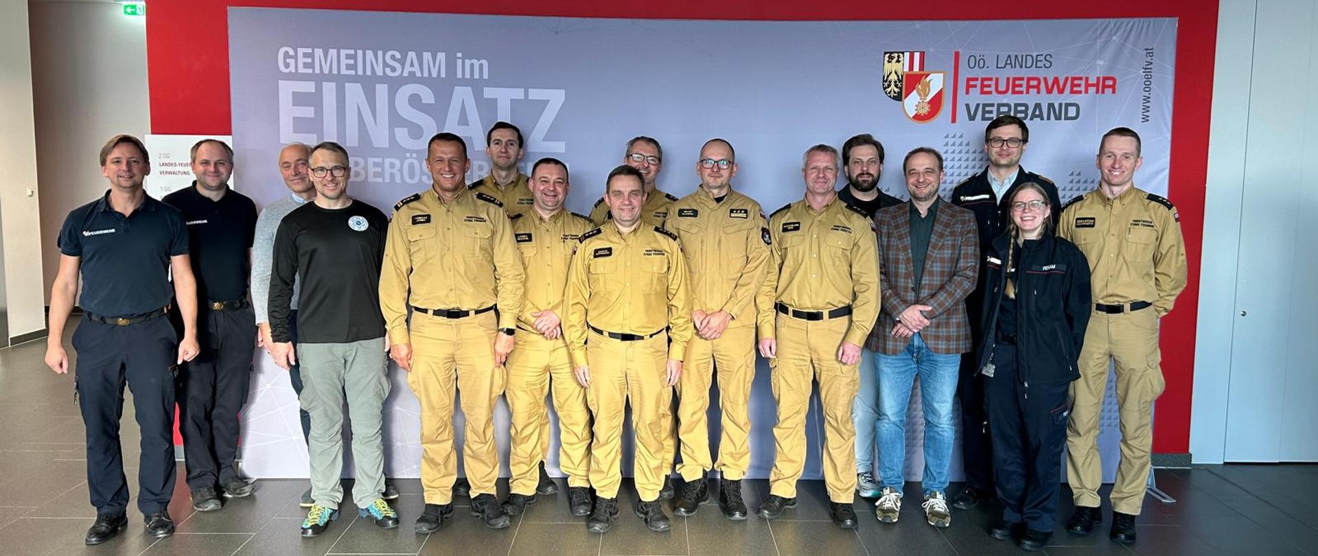 Grupa około dwudziestu osób, w tym strażacy PSP w beżowych mundurach oraz kilku cywilów, stoi wspólnie na tle ścianki z napisem „Gemeinsam im Einsatz” i logo Feuerwehrverband Oberösterreich. Wszyscy pozują do zdjęcia grupowego w jasnym holu budynku.