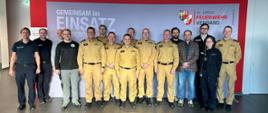 Grupa około dwudziestu osób, w tym strażacy PSP w beżowych mundurach oraz kilku cywilów, stoi wspólnie na tle ścianki z napisem „Gemeinsam im Einsatz” i logo Feuerwehrverband Oberösterreich. Wszyscy pozują do zdjęcia grupowego w jasnym holu budynku.