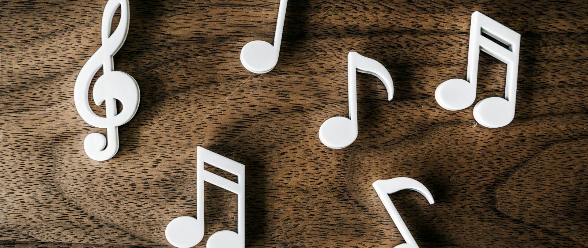 grafika przedstawiająca biały klucz wiolinowy oraz nuty ułożone na ciemnym, drewnianym blacie