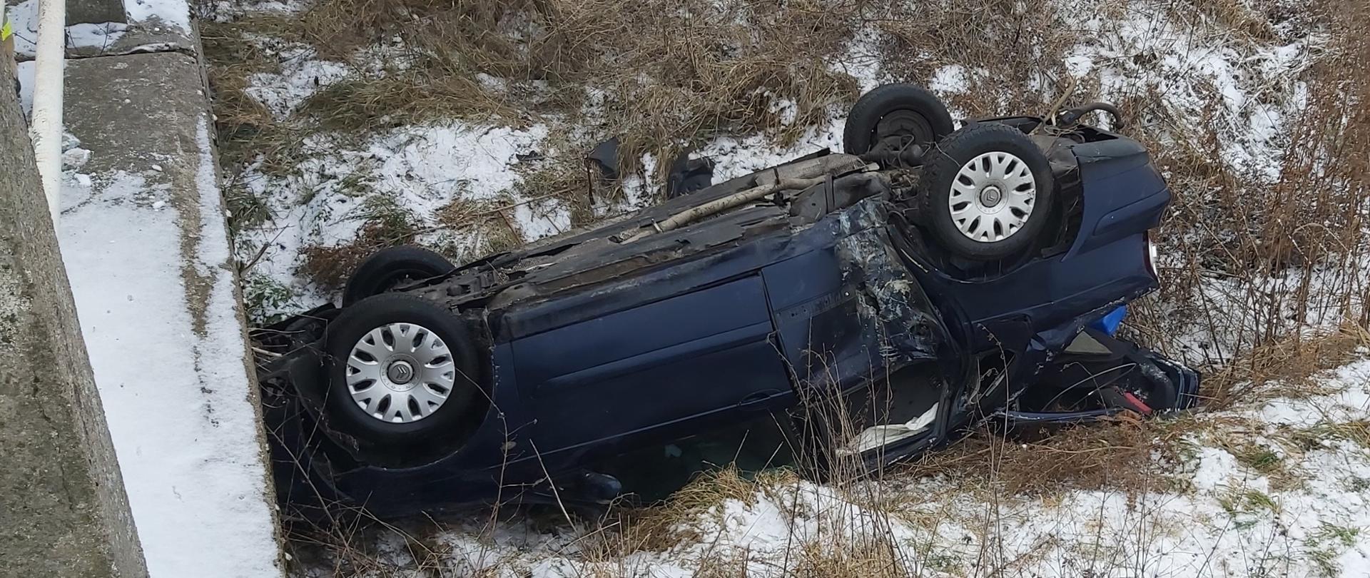 Auto osobowe leży na dachu obok betonowego przepusty w przydrożnym rowie. Tylne drzwi od strony pasażera uszkodzone po uderzeniu w mostek. Teren wokół pokryty niewielką warstwą śniegu.