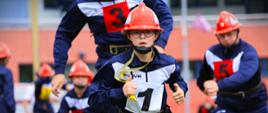 XII Wojewódzkie Młodzieżowe Zawody Sportowo-Pożarnicze Białystok 2025 - zawodnicy w trakcie rywalizacji - chłopiec w granatowym dresie, w pasie strażackim i kasku biegnie, trzymając żółty gwizdem, za nim inni chłopy z drużyny 