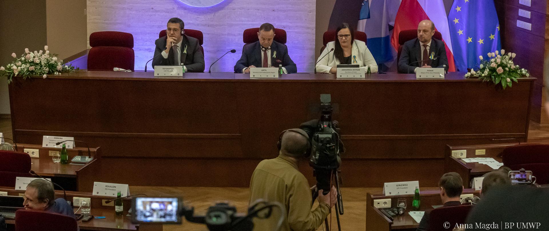 Przy długim stole siedzą cztery osoby. Druga od prawej wiceminister Małgorzata Jarosińska-Jedynak. Za nimi flagi PL, UE i UA. Na ścianie godło Rzeszowa. Przed nimi kamerzysta i słuchacze.