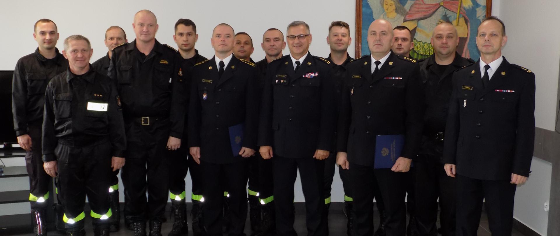 Obraz przedstawia grupę strażaków znajdującą się w świetlicy Komendy Powiatowej Państwowej Straży Pożarnej w Rypinie. W środku pierwszego rzędu od lewej: mł. asp. Rafał Raczkowski, st. bryg. Jacek Kaczmarek Kujawsko-Pomorski Komendant Wojewódzki, st. bryg. Grzegorz Łydkowski Komendant Powiatowy PSP w Rypinie. W tle obraz św. Floriana