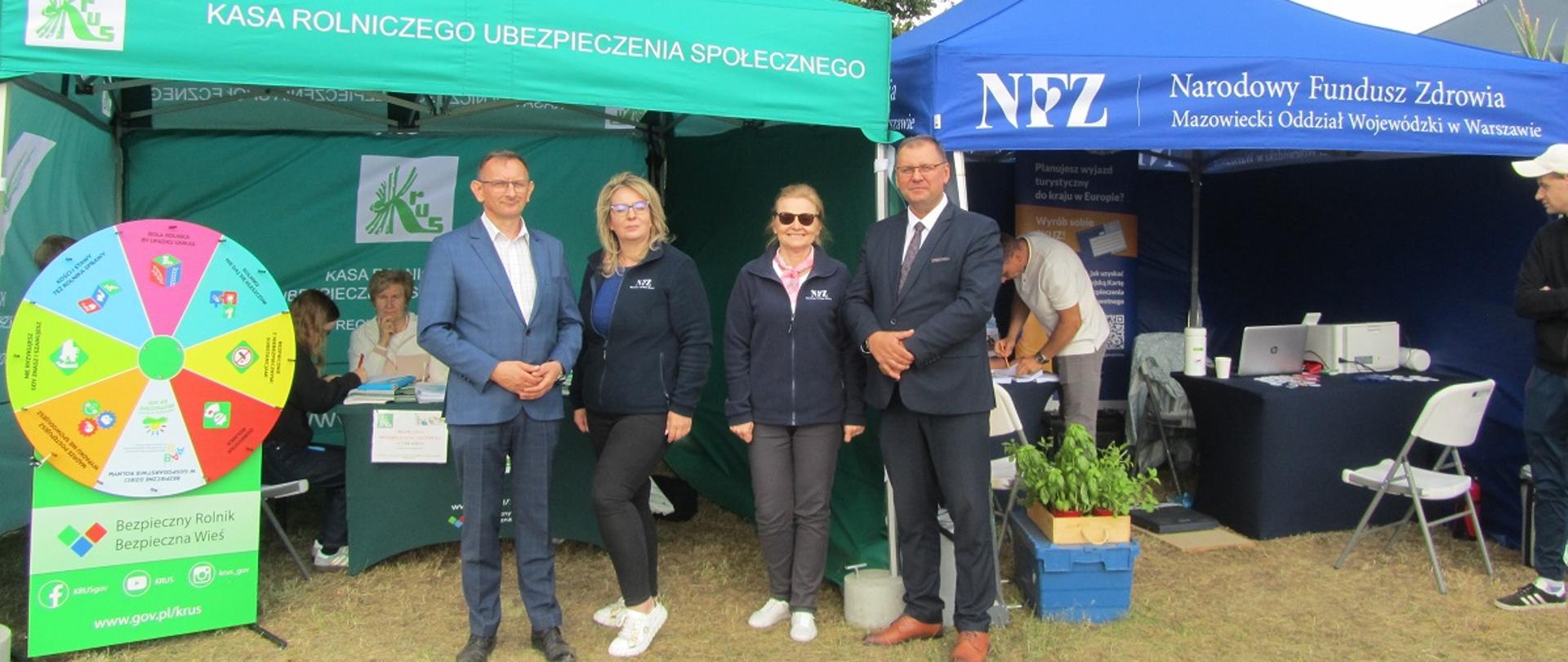 Grupa pięciu osób stojących przed dwoma namiotami wystawowymi. Po lewej stronie znajduje się zielony namiot z logo i napisem „Kasa Rolniczego Ubezpieczenia Społecznego”, a obok kolorowe koło fortuny. Po prawej stronie stoi ciemnoniebieski namiot z napisem „NFZ – Narodowy Fundusz Zdrowia, Mazowiecki Oddział Wojewódzki w Warszawie”. Osoby stojące na środku uśmiechają się do zdjęcia, dwie Panie ubrane są w granatowe polary z logo NFZ, a dwaj mężczyźni po bokach mają garnitury. W tle widać osoby siedzące przy stołach wewnątrz namiotów. Namioty stoją na trawiastym terenie.