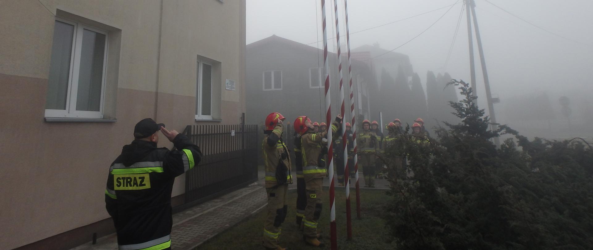 Przed budynkiem komendy PSP trójka strażaków ubranych w ubrania specjalne stoją przy maszcie flagowym i wciągają flagę państwową na maszt. Za nimi stoją na baczność w dwóch rzędach pozostali strażacy również w strojach specjalnych.