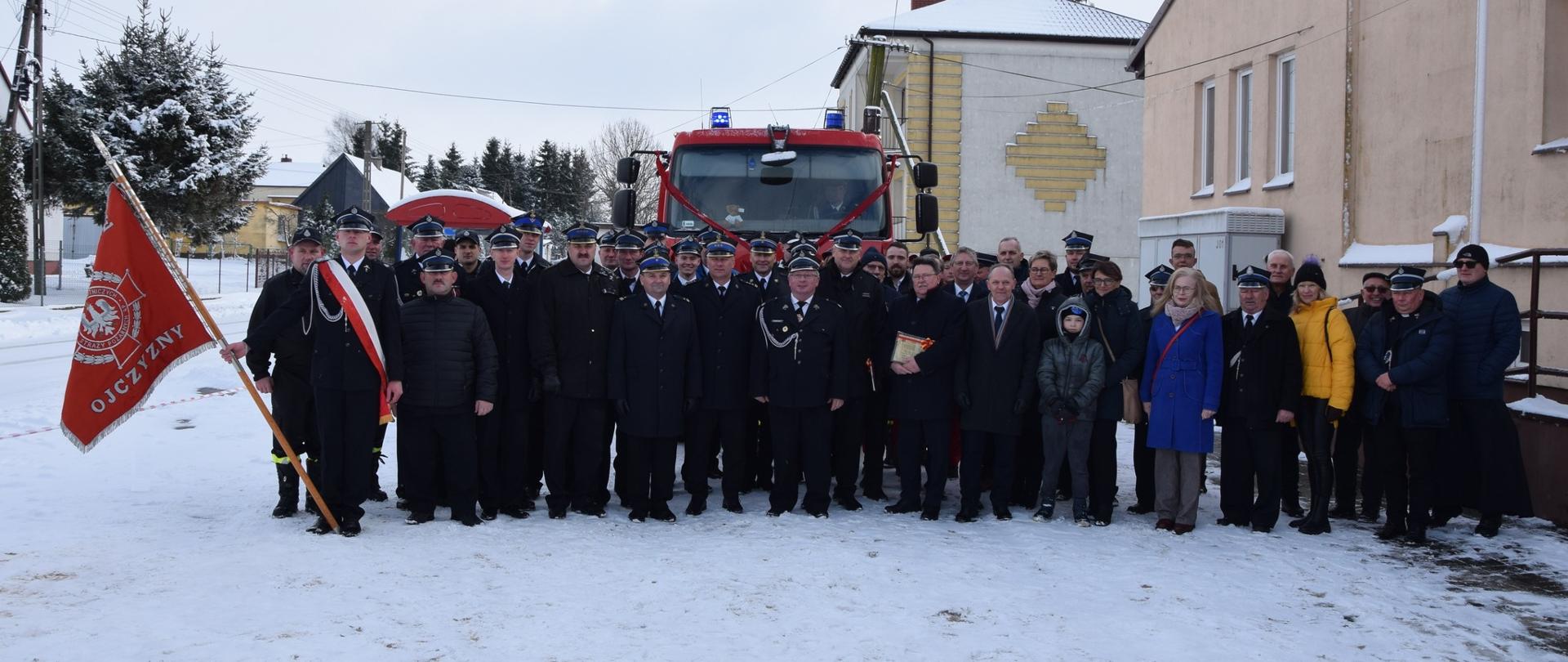 Przed przekazanym samochodem ratowniczo-gaśniczym stoją strażacy i goście - uczestniczy uroczystej zbiórki w miejscowości Dulsk.