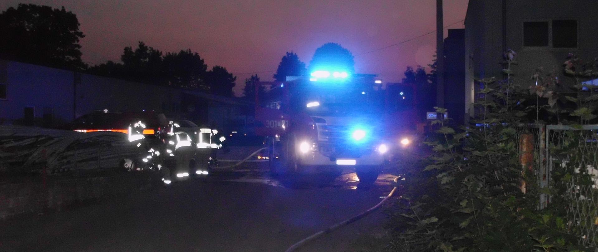 Zdjęcie przedstawia teren akcji - widać rozwiniętą linię gaśniczą. W tle znajdują się samochody pożarnicze oraz strażacy w odblaskowych ubraniach specjalnych. Na zewnątrz panuje półmrok. Nie widać zadymienia.