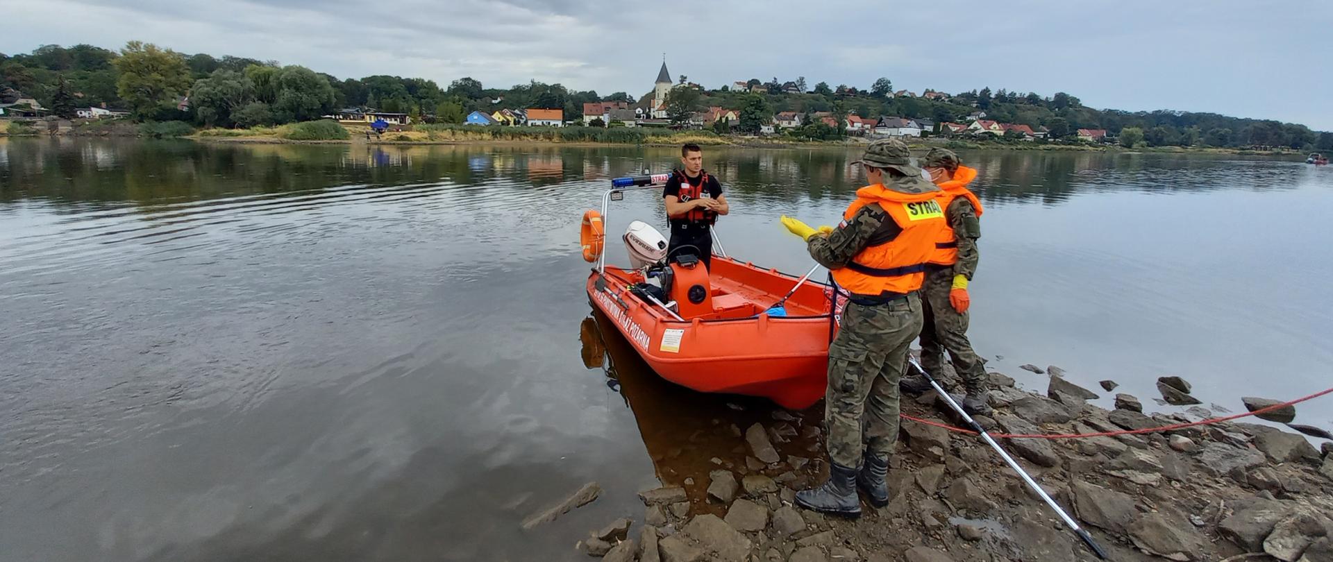 Strażak w kapoku w łódce na brzegu rzeki. Przed łodzią stojący dwaj żołnierze ubrani w kapoki.