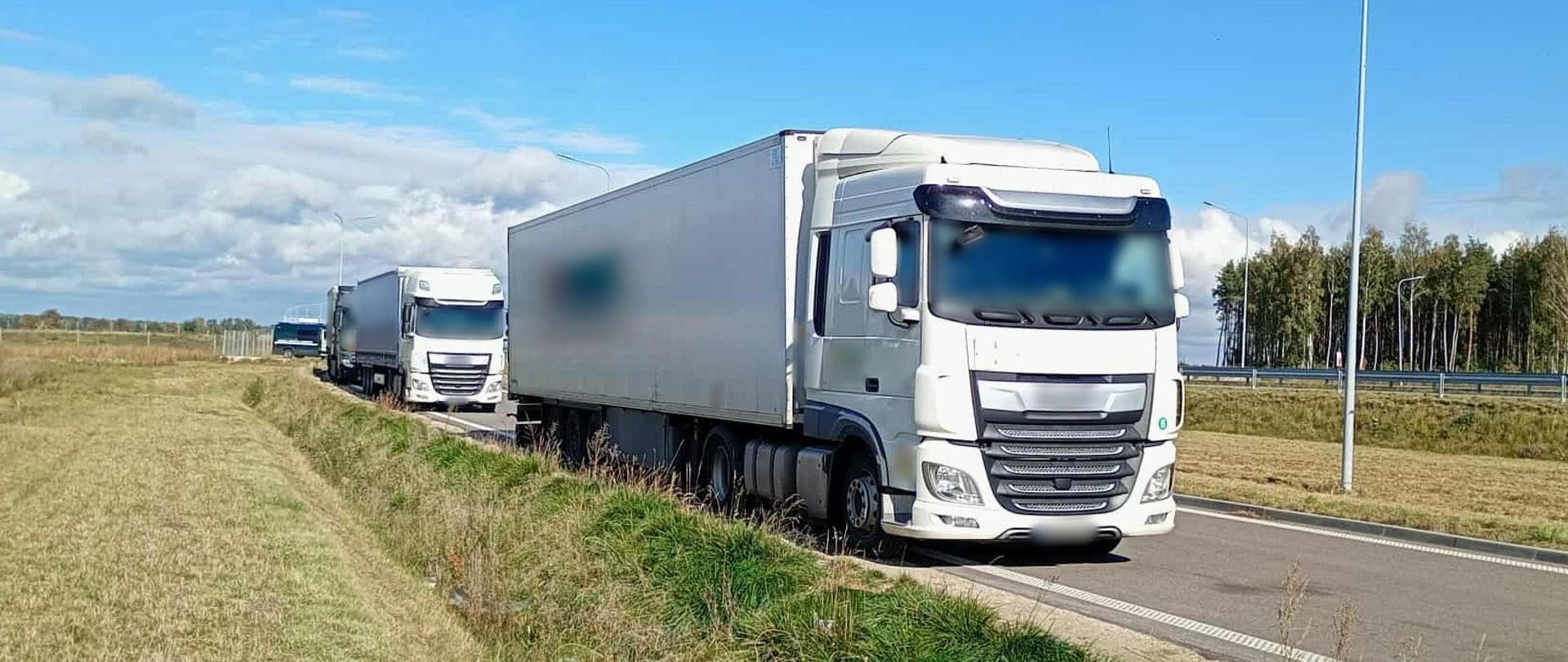 Na pierwszym planie: trzy zatrzymane do kontroli zestawy ciężarowe stoją w wyznaczonym miejscy przy drodze ekspresowej S61 pod Ostrowią Mazowiecką. W tle: za kontrolowanymi ciężarówkami stoi zaparkowany w poprzek oznakowany furgon mazowieckiej Inspekcji Transportu Drogowego.