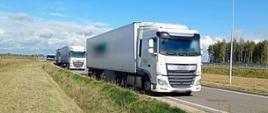 Na pierwszym planie: trzy zatrzymane do kontroli zestawy ciężarowe stoją w wyznaczonym miejscy przy drodze ekspresowej S61 pod Ostrowią Mazowiecką. W tle: za kontrolowanymi ciężarówkami stoi zaparkowany w poprzek oznakowany furgon mazowieckiej Inspekcji Transportu Drogowego.