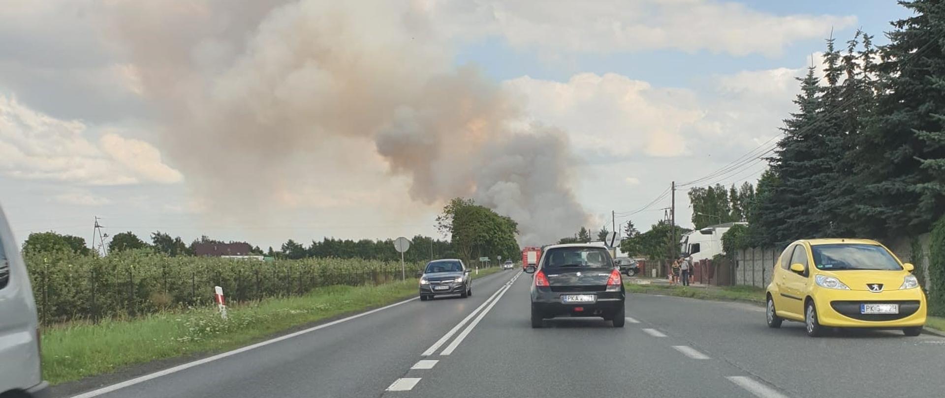 Na zdjęciu widać Drogę Krajową nr 12. W obu kierunkach przemieszczają się samochody. Po bokach drogi drzewa, korony zielone. Droga posiada oznakowanie poziome w kolorze białym. W centralnym punkcie duże zadymienie, dym koloru szarego. Na niebie widoczne chmury.
