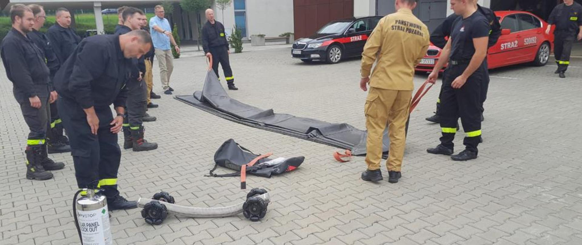 Strażacy na placu wewnętrznym Komendy Miejskiej Państwowej Straży Pożarnej w Mysłowicach testują płachtę gaśniczą do gaszenia samochodów w tym samochodów elektrycznych oraz rolki do transportu węży pożarniczych.