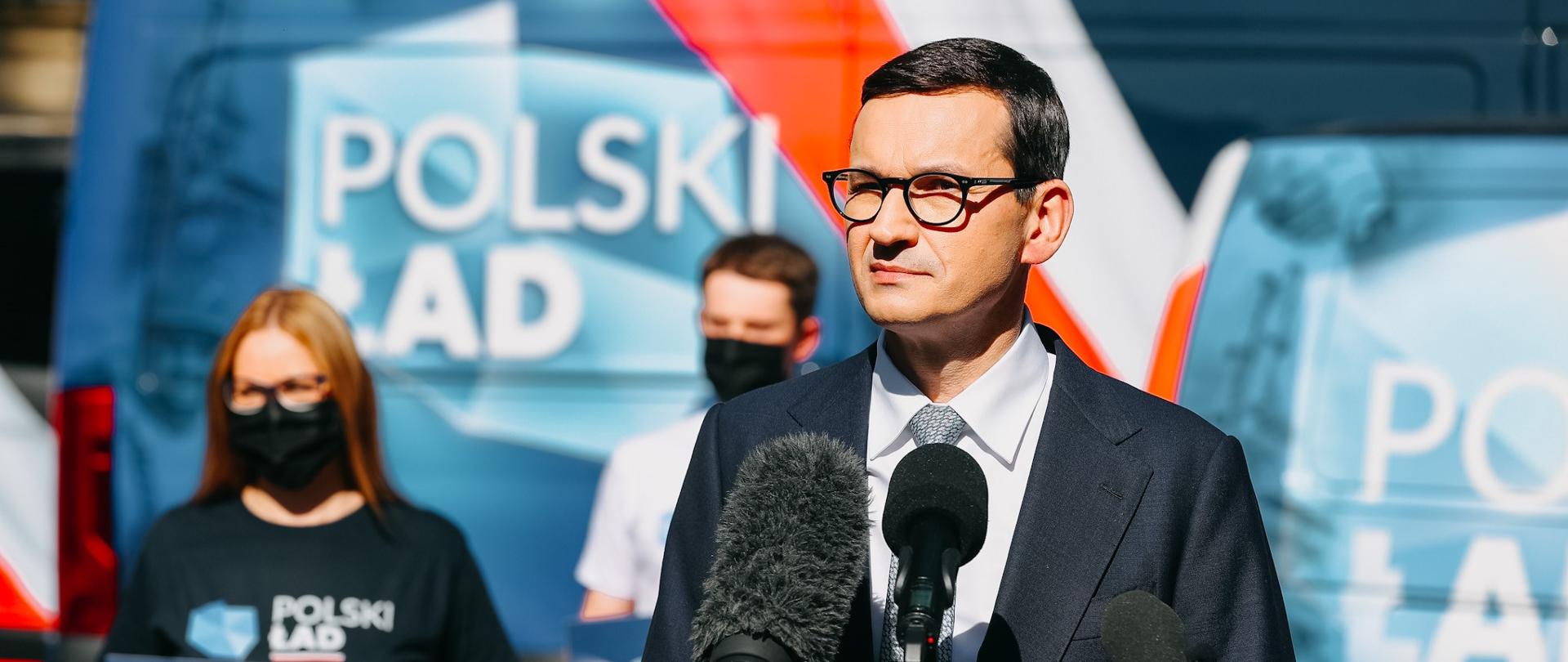 Premier Mateusz Morawiecki stoi przy mikrofonie, w tle samochody z napisem Polski Ład.