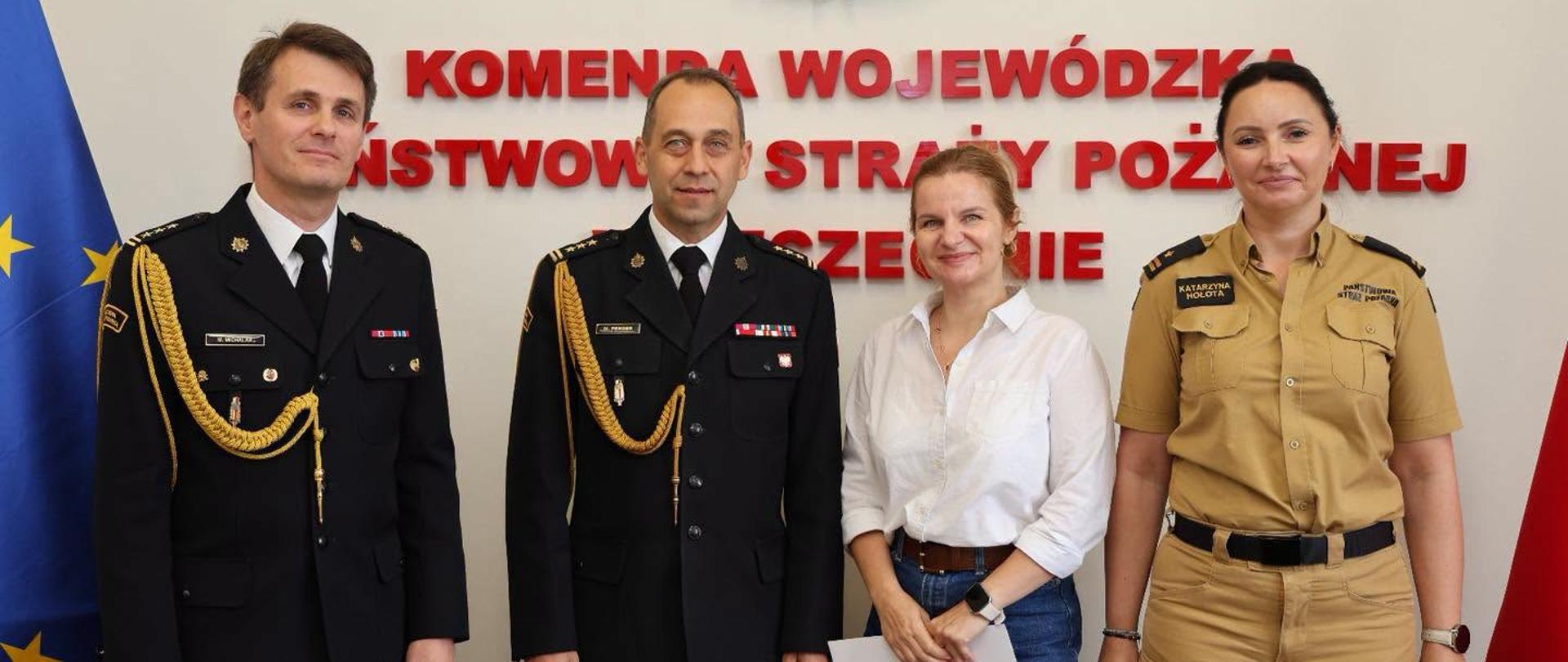 Do wspólnego zdjęcia w holu Komendy Wojewódzkiej PSP w Szczecinie pozują dwaj funkcjonariusze Państwowej Straży Pożarnej w galowych mundurach, kobieta w białej koszuli oraz funkcjonariuszka PSP w piaskowym mundurze służbowym. W tle znajdują się godło Państwowej Straży Pożarnej, czerwony napis z nazwą komendy oraz flagi Polski i Unii Europejskiej.