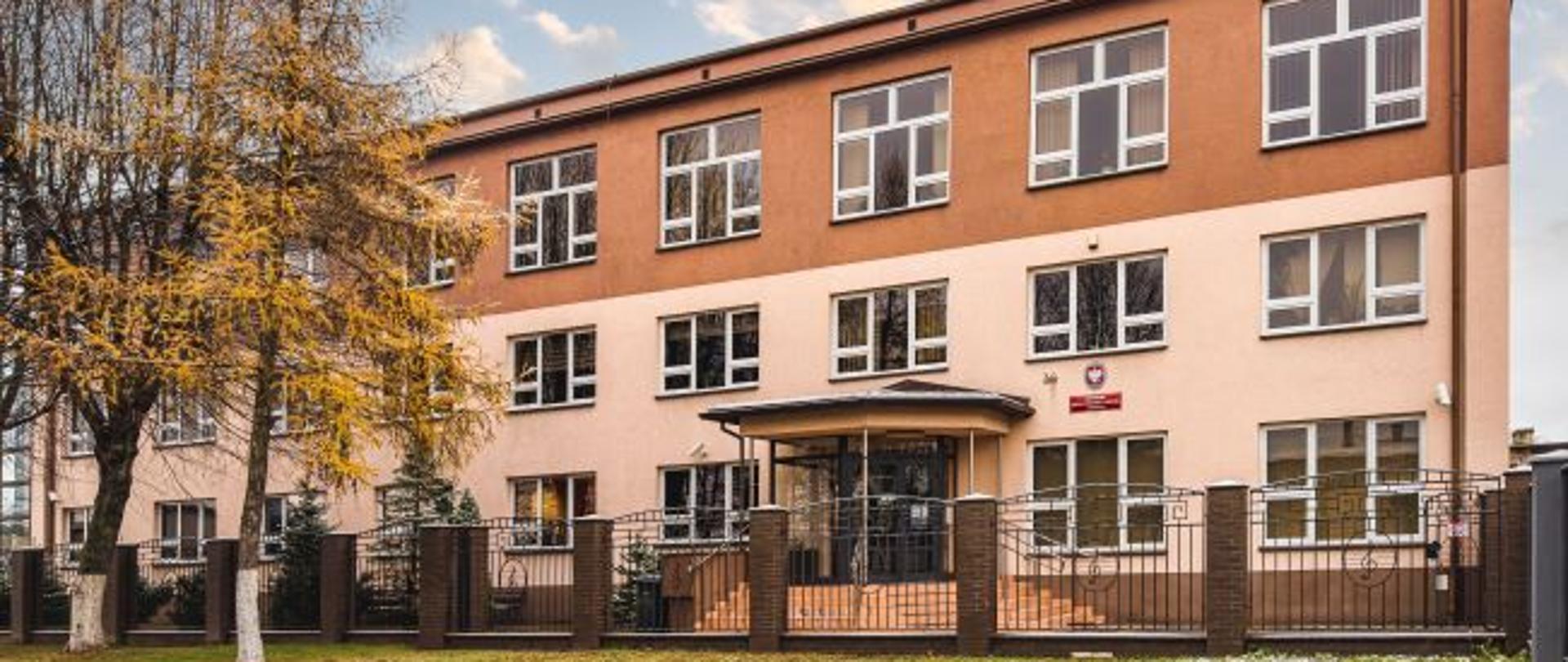 Zdjęcie przedstawia budynek szkoły muzycznej w Kolbuszowej od strony frontowej. Dwupiętrowy budynek jest koloru beżowo-brązowego. Do wejścia głównego szkoły prowadzą kilkustopniowe schody. Szkoła ogrodzona jest płotem z elementami metalowymi oraz słupkami.. Po lewej stronie widać drzewa.