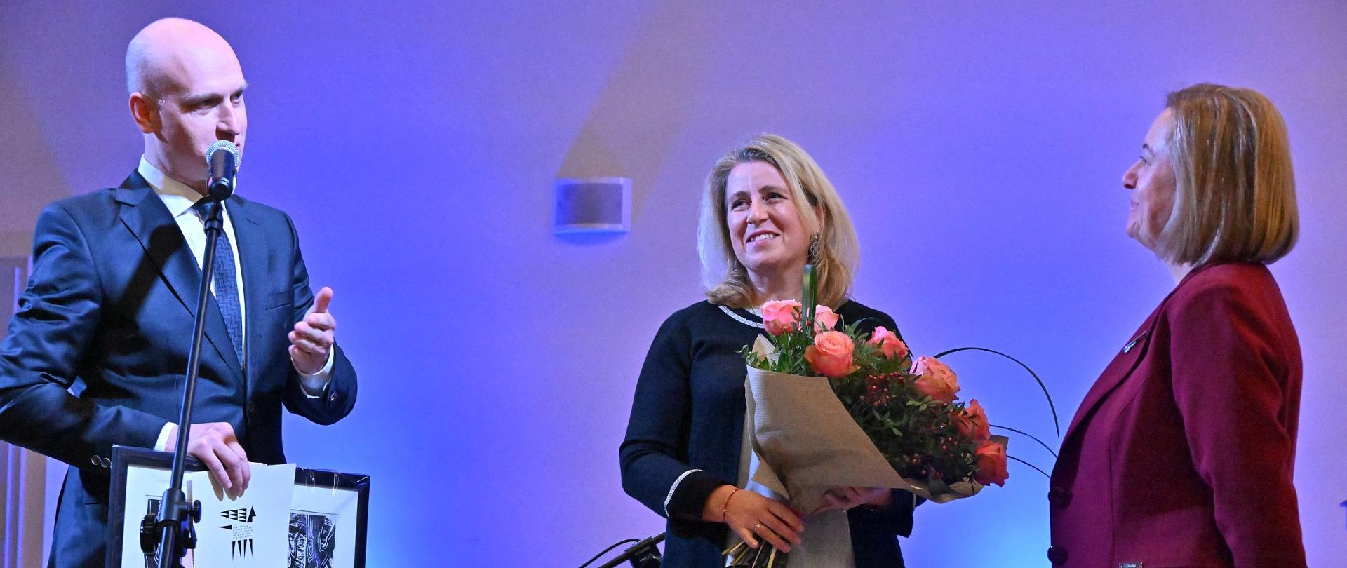 Na zdjęciu od lewej strony znajduje się Marek Król-Józaga dyrektor Państwowego Liceum Sztuk Plastycznych im. Antoniego Kenara w Zakopanem. Dyrektor trzymając w ręku prezent - grafikę autorstwa Pauliny Wysoczyńskiej uczennicy kl. 4C składa gratulacje z okazji jubileuszu 30-lecia szkoły pani Zofie Ziach dyrektorowi Państwowej Ogólnokształcącej Szkoły Artystycznej stojącej po prawej stronie. W środku z kwiatami stoi pani Agnieszka Wójcik-Marduła wicedyrektor PLSP im. A. Kenara w Zakopanem