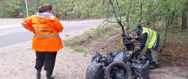 Dwójka inspektorów WIOŚ w Warszawie porządkuje worki ze śmieciami na poboczu leśnej, asfaltowej drogi. Z worków wystają plastikowe butelki i opakowania. Pośród worków leży gumowa opona.