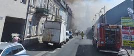 Zdjęcie przedstawia pożar kamienicy. Na zdjęciu widać kamienice w zabudowie pierzejowej. Dym unosi się nad budynkiem. oraz częściowo nad ulicą. Ponadto na zdjęciu widać samochody strażackie. Na ulicy leżą linie wężowe. 