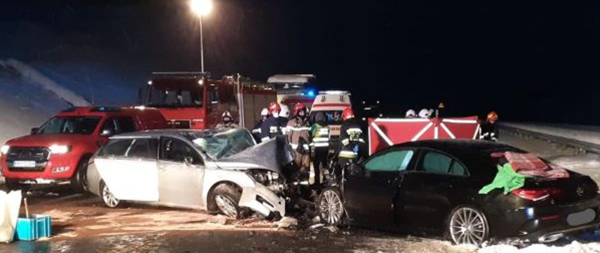 Zdjęcie przedstawia w środkowej części rozbite samochody osobowe po zderzeniu czołowym, grupę strażaków oraz na drugim planie samochody pożarnicze i karetki pogotowia ratunkowego. Zdjęcie zrobione w porze nocnej, teren akcji oświetlony z najaśnic samochodów pożarniczych.