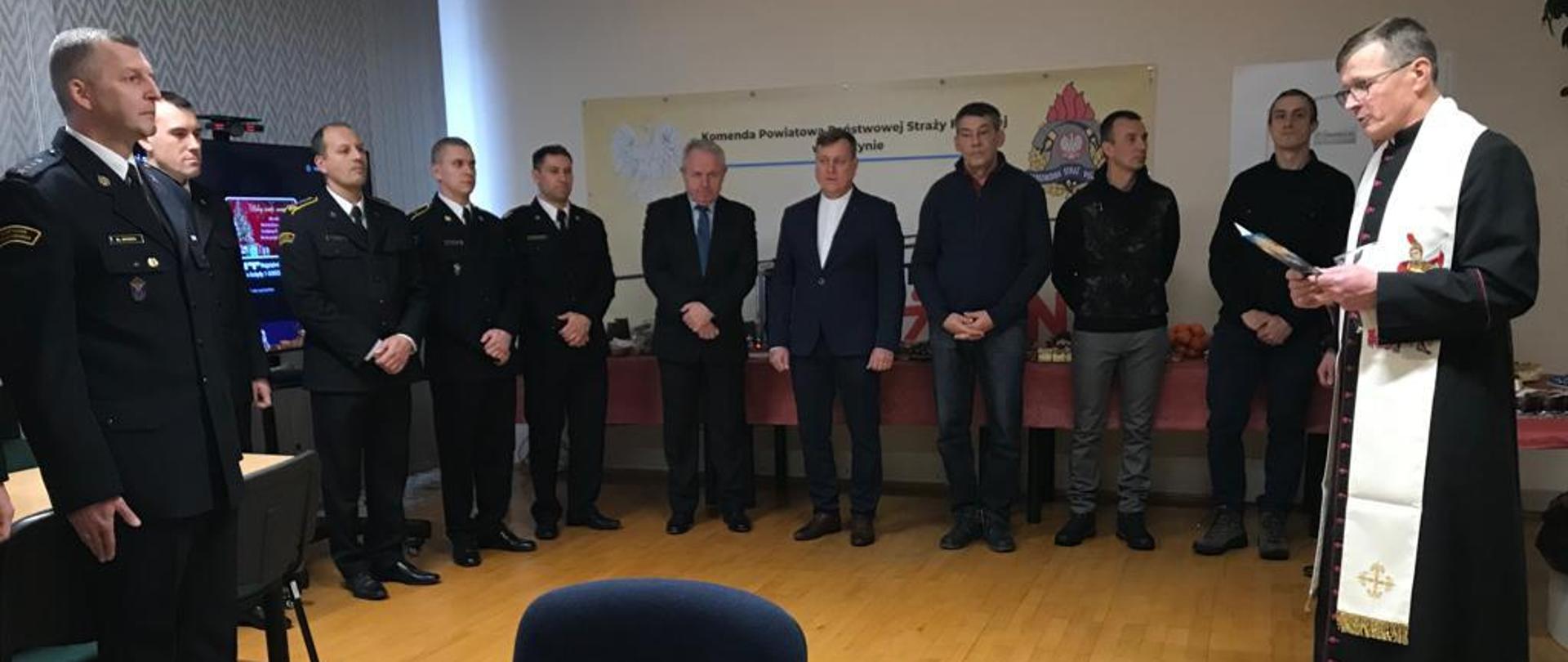 Zdjęcie zrobione Sali Konferencyjnej kętrzyńskiej komendy. Funkcjonariusze ubrani w czarne mundury wyjściowe. Ksiądz odmawia modlitwę, po prawej stronie stoi Starosta Kętrzyński oraz Komendant Powiatowy.