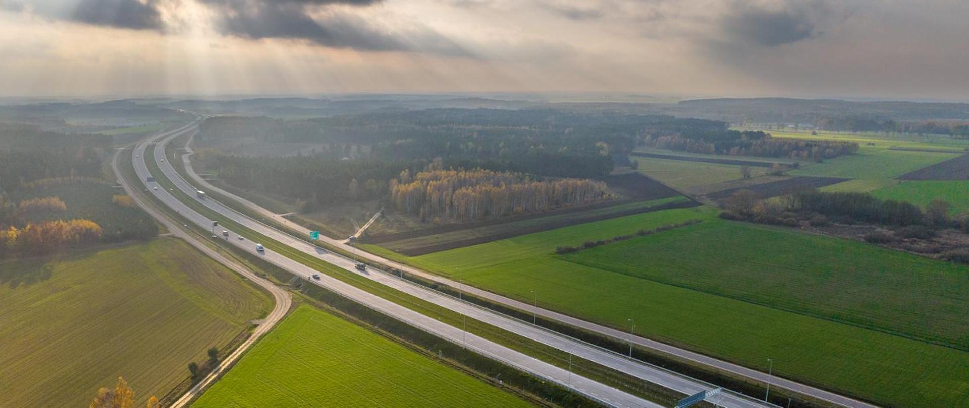Droga szybkiego ruchu, wokół niej pola i lasy, zza chmur przebija się słońce, zdjęcie lotnicze