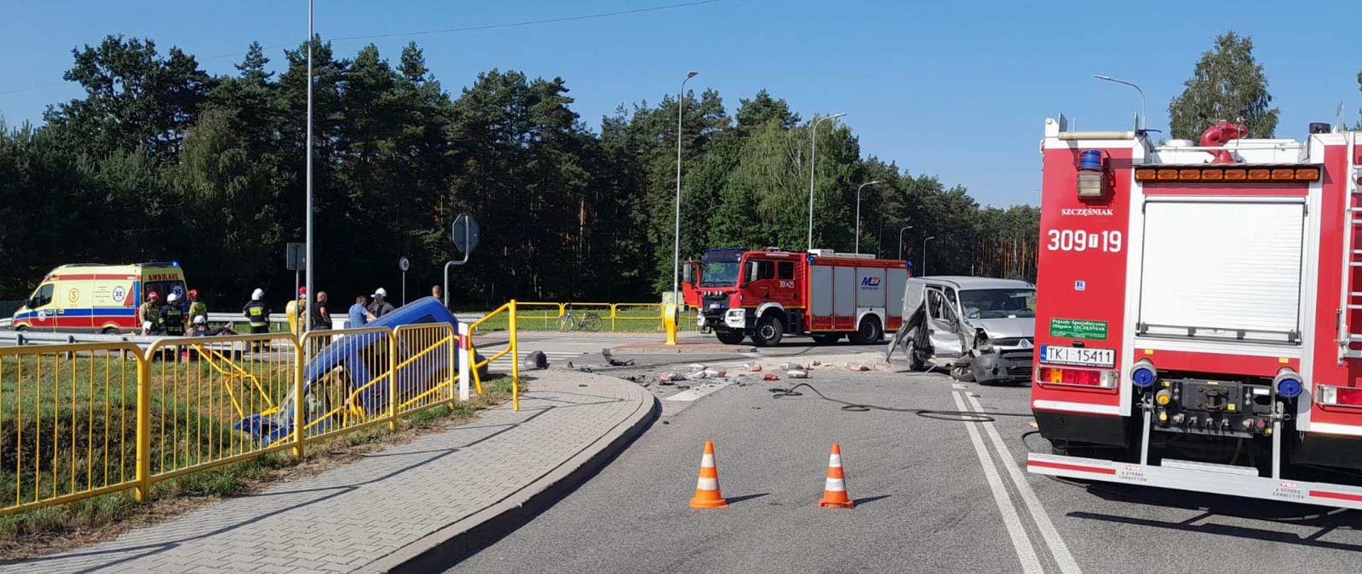 Zdjęcie przedstawia skrzyżowanie gdzie doszło do zderzenia. Od prawej stoją: samochód pożarniczy, pierwszy z uszkodzonych aut, drugi wóz strażacki, drugi z uszkodzonych aut (pochylony do przodu znajduje się w przydrożnym rowie) i ambulans Państwowego Pogotowia Ratunkowego. 