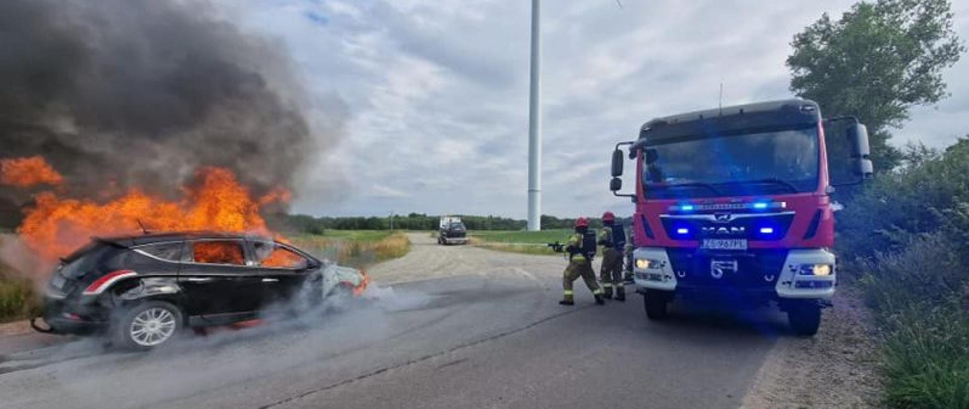Zdjęcie przedstawia moment dojazdu służb do palącego się pojazdu. Auto całkowicie objęte ogniem. Do auta dobiegaj strażacy z odciekiem. Zdjęcie uchwyciło moment rozpoczęcia działań. Widać na nim również pojazd gaśniczy.