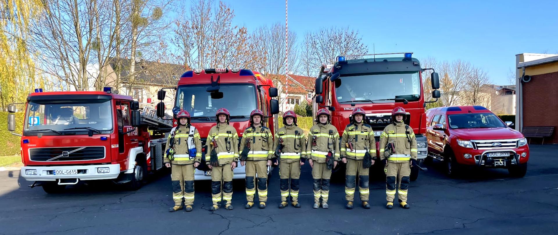 Na zdjęciu strażacy stoją w szeregu w ubraniach specjalnych koloru beżowego i czerwonych hełmach, z tył samochody pożarnicze oraz maszt z Flagą Polski.