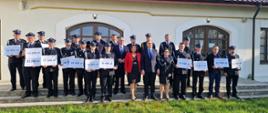 Strażacy ochotnicy stoją na trzech schodkach przed budynkiem trzymając w dłoniach tabliczki z kwotami udzielonych dotacji. Razem z nimi stoją Prezes Zarządu Wojewódzkiego Funduszu Ochrony Środowiska i Gospodarki Wodnej w Warszawie Pan Marek Ryszka, Sekretarz Stanu w Ministerstwie Aktywów Państwowych Pan Maciej Małecki, Komendant Powiatowy Państwowej Straży Pożarnej w Żyrardowie st. bryg. Mariusz Tymoszewicz oraz Wójt Gminy Radziejowice Pani Urszula Ciężka.