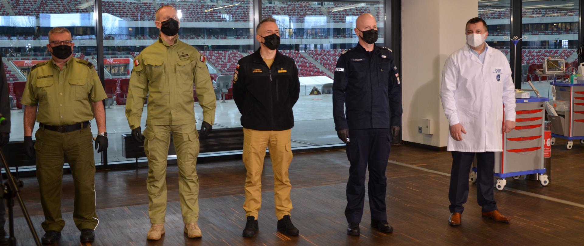 Przedstawiciele służb mundurowych w mundurach, w tle trybuna stadionu narodowego w Warszawie. Po ich prawej stronie stoi lekarz.