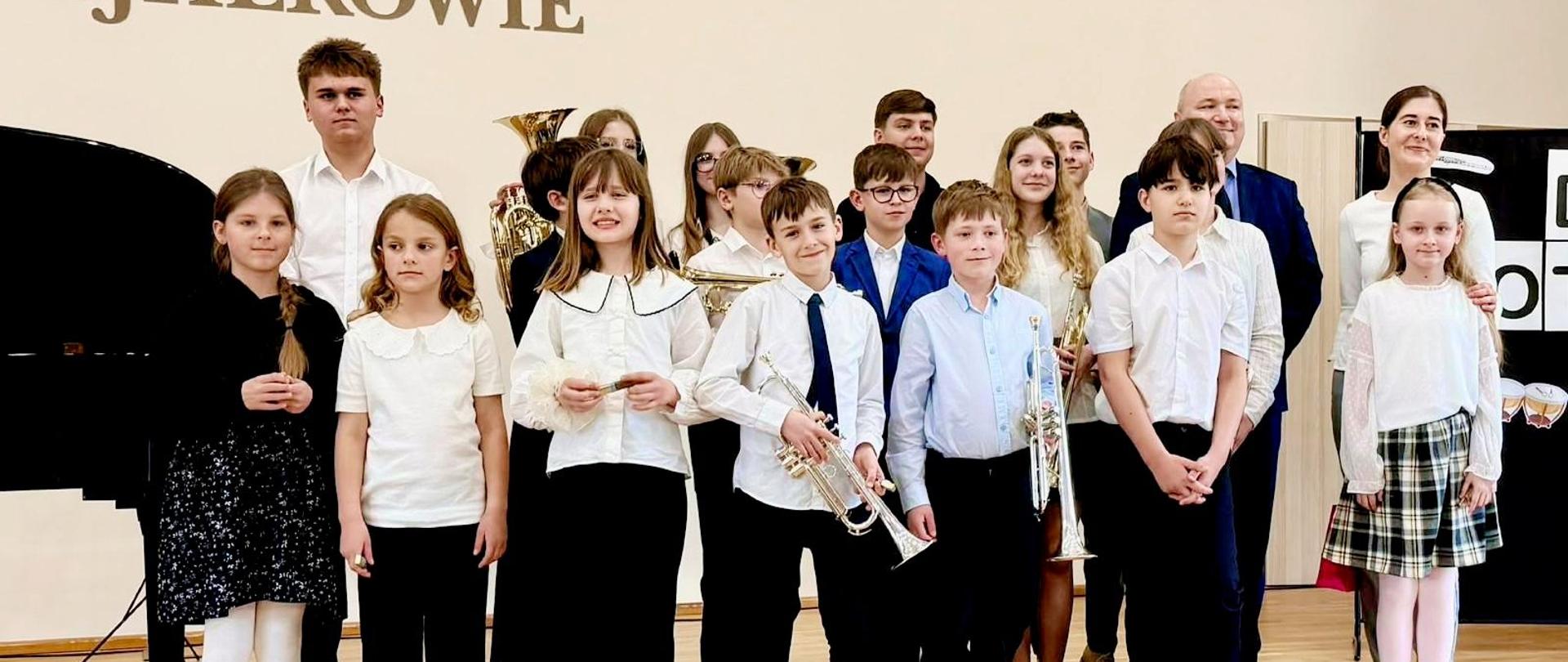 Na scenie sali koncertowej PSM pozują uczniowie biorący udział w koncercie wraz z nauczycielami.