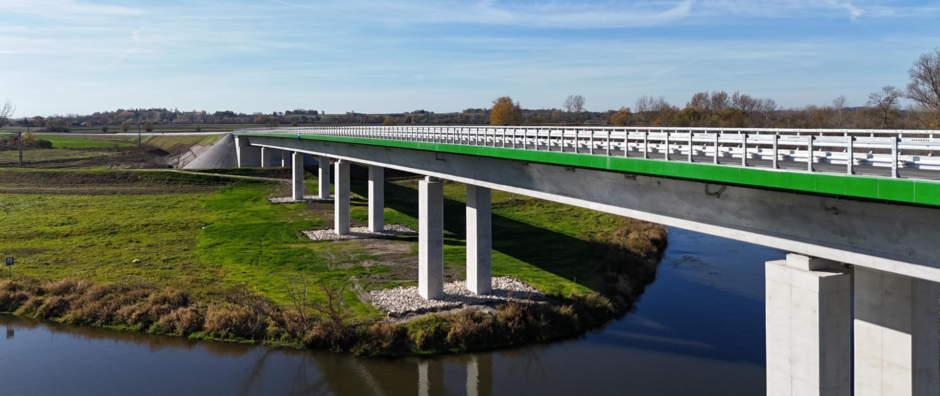 Nowoczesny most drogowy na betonowych filarach nad rzeką, otoczony zielonymi łąkami; jasne niebo i wiejski krajobraz w tle.