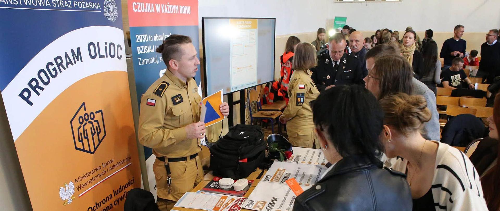 Strażak Państwowej Straży Pożarnej st. asp. Mikołaj Wójcik prezentuje tabliczkę z międzynarodowym znakiem obrony cywilnej zgromadzonym odwiedzającym przy stoisku edukacyjno-profilaktycznym.