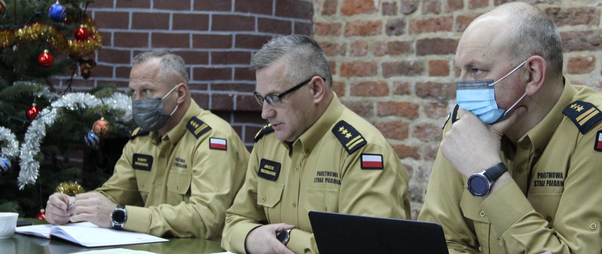 Przy stole prezydialnym podczas narady siedzą komendanci - wojewódzkie PSP i jego zastępcy. Są w umundurowaniach służbowych, na twarzach maseczki ochronne. W tle wystrój świąteczny. Trwa narada. Na stole materiały - notatniki, komputer...