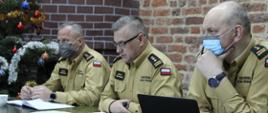 Przy stole prezydialnym podczas narady siedzą komendanci - wojewódzkie PSP i jego zastępcy. Są w umundurowaniach służbowych, na twarzach maseczki ochronne. W tle wystrój świąteczny. Trwa narada. Na stole materiały - notatniki, komputer...