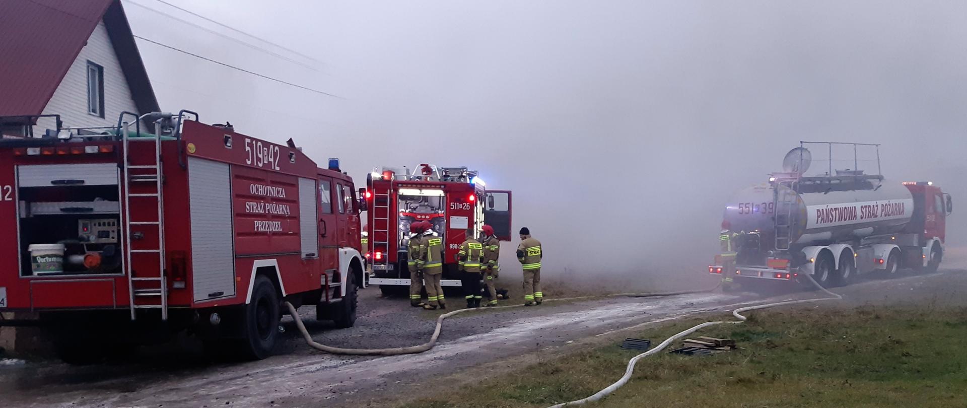 dwa czerwone samochody strażackie oraz samochód cysterna i strażacy w ubraniach specjalnych na tle zadymionego pogorzeliska. Rozwinięte węże pożarnicze pomiędzy pojazdami