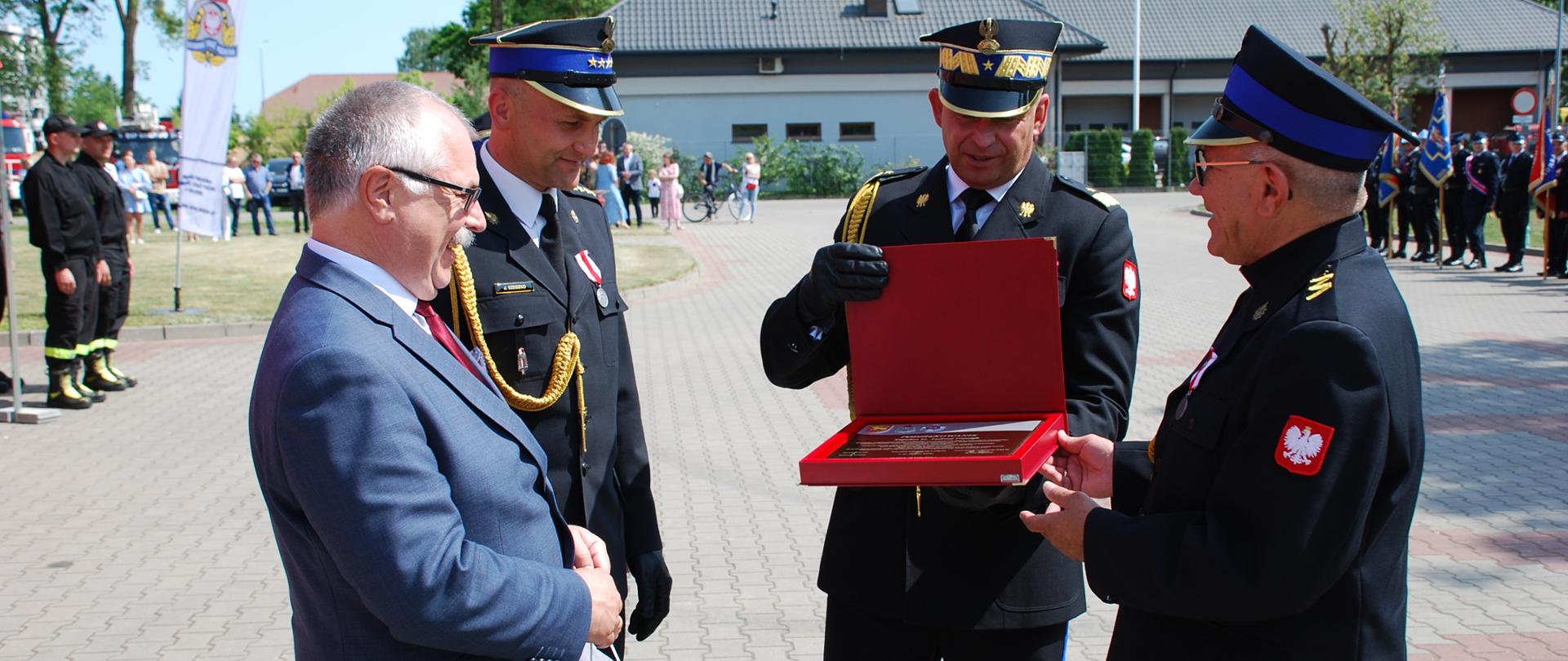 Funkcjonariusze PSP w mundurach wyjściowych oraz mężczyzna w garniturze wręczają pamiątkowy grawerton umundurowanemu księdzu kapelanowi