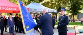 Pani starosta lęborski przekazuje sztandar wicewojewodzie pomorskiemu w obecności zaproszonych gości. Obok stoi strażak z mikrofonem.