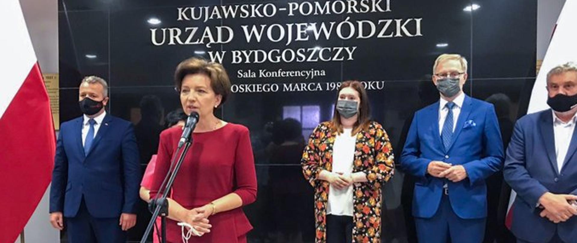 Zdjęcie grupowe. Na pierwszym planie stoi przy mikrofonie ubrana w czerwony kostium minister Maląg. Z tyłu stoja trzej mężczyźni i jedna kobieta. Za ich plecami napis Kujawsko-Pomorski Urząd Wojewódzki w Bydgoszczy.