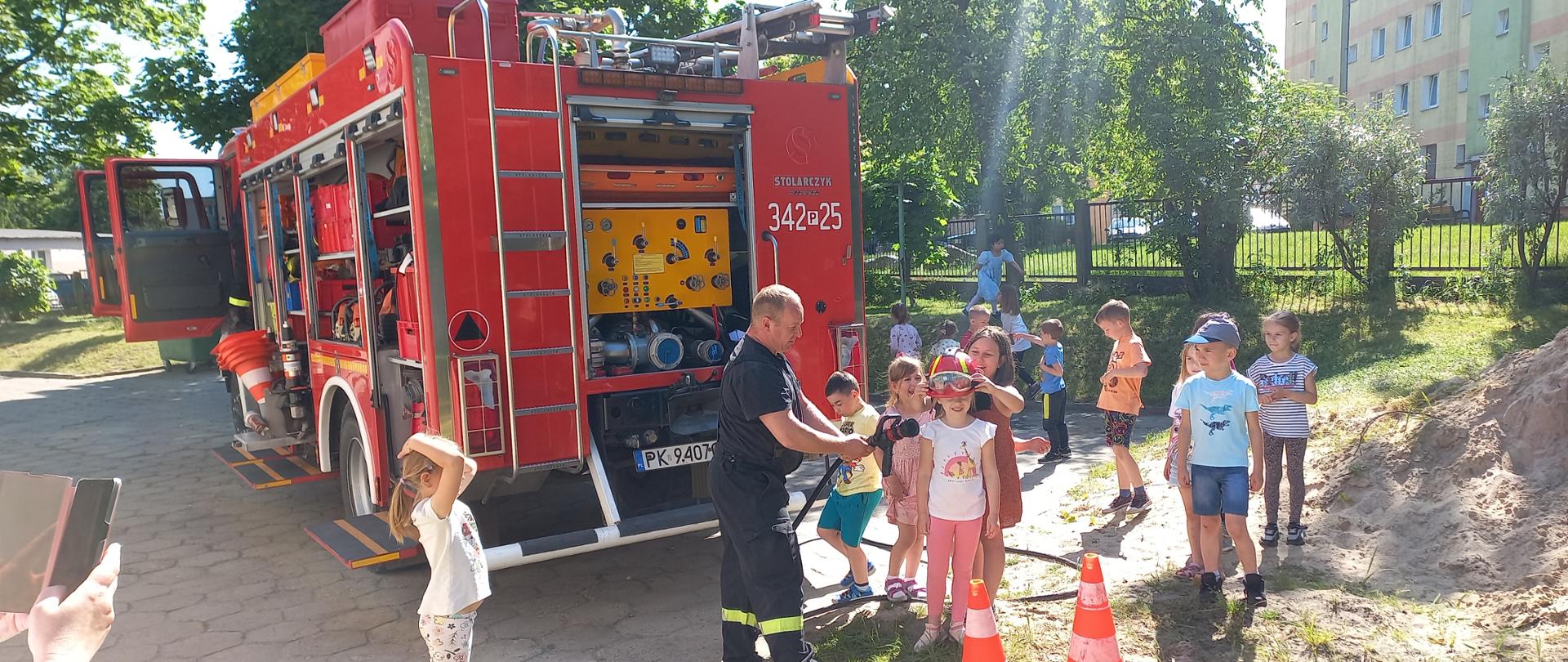 Zdjęcie wykonane na terenie przedszkola. Widać trawę oraz drzewa. W centrum zdjęcia strażak oraz dziecko. Widać zabudowę samochodu ratowniczo-gaśniczego.