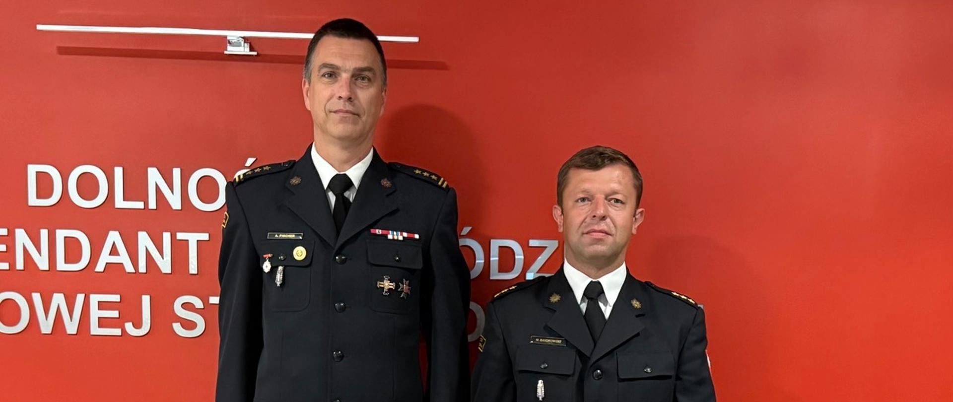 Strażacy w ciemnych mundurach wyjściowych stoą na tle czerwonej ściany z białym napisem „DOLNOŚLĄSKA KOMENDA WOJEWÓDZKA PAŃSTWOWEJ...”. Mężczyzna trzyma czerwoną teczkę z godłem Polski.