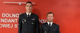Strażacy w ciemnych mundurach wyjściowych stoą na tle czerwonej ściany z białym napisem „DOLNOŚLĄSKA KOMENDA WOJEWÓDZKA PAŃSTWOWEJ...”. Mężczyzna trzyma czerwoną teczkę z godłem Polski.