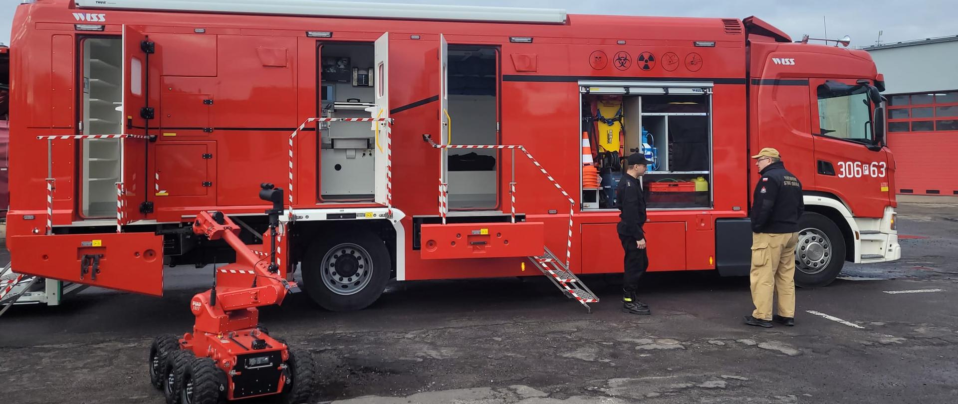 duży, czerwony samochód strażacki, przed nim stoją strażacy, obok robot na kółkach 