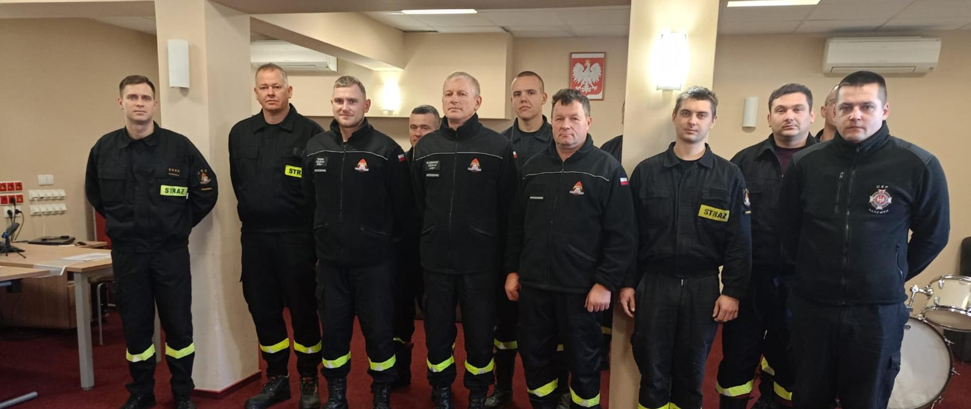W pomieszczeniu na czerwonym dywanie w dwuszeregu stoi kilkunastu strażaków w mundurach.