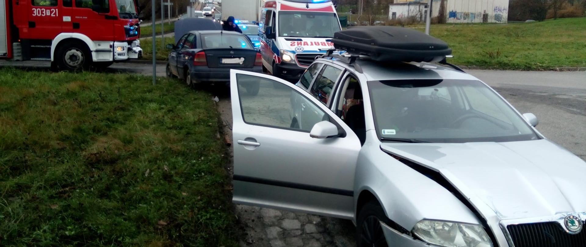 Zdjęcie przedstawia oba samochody biorące udział w wypadku. Za nimi, po prawej stoi ambulans a po lewej samochód pożarniczy.