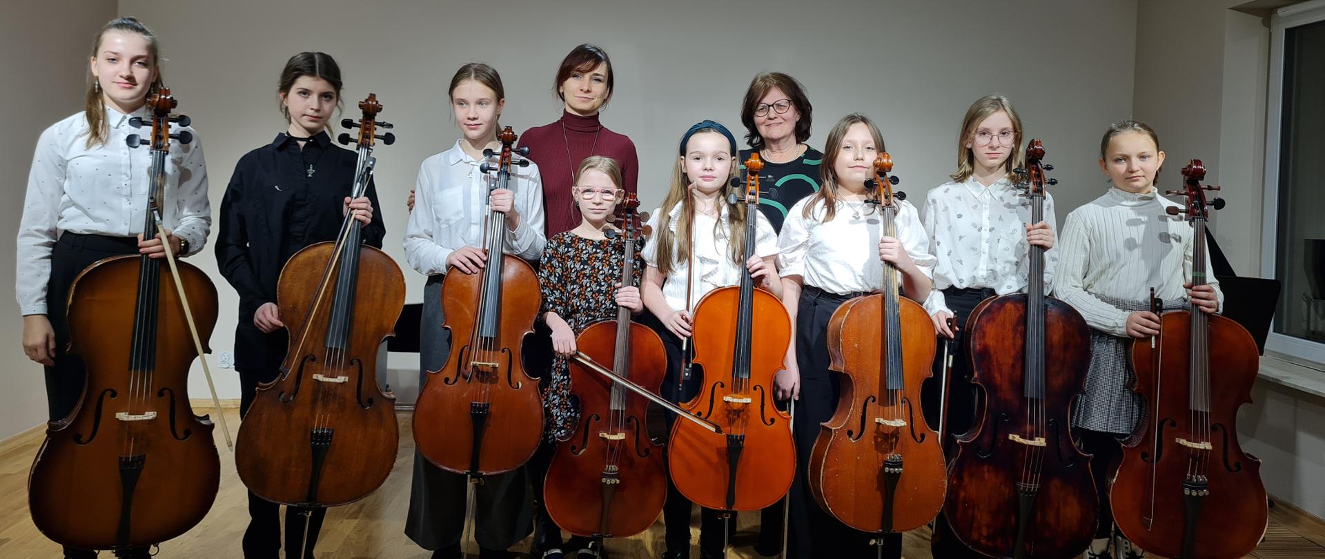 Zdjęcie przedstawia grupę uczennic stojących na scenie sali kameralnej, trzymających w ręku wiolonczele. Uczennicom towarzyszą ich nauczycielki.