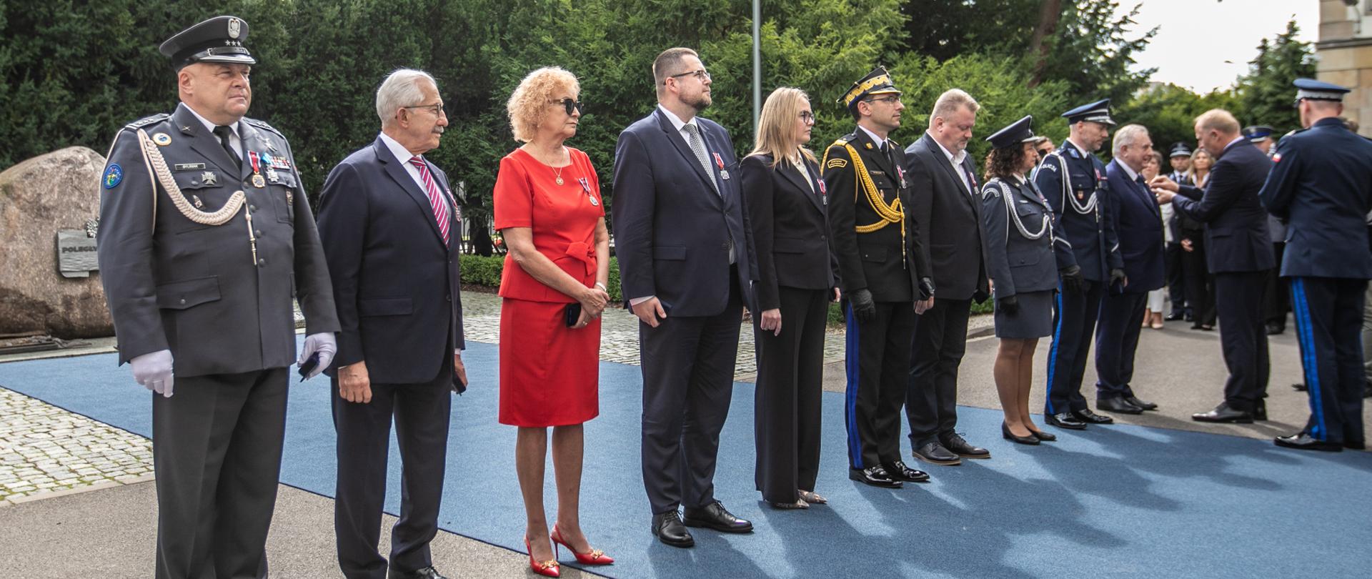 Szereg odznaczonych funkcjonariuszy oraz gości honorowych stojących w kolejce podczas uroczystości przed Komendą Główną Policji. Na pierwszym planie widoczny oficer w szarym mundurze galowym, obok osoby cywilne i funkcjonariusze PSP oraz Policji.