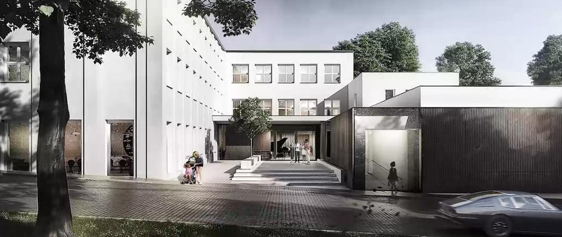 Zdjęcie przedstawia widok dwupiętrowego, białego budynku szkoły znajdującego się za wyłożoną kostką brukową drogą. Z przodu budynku znajduje się przeszkolone wejście, przed którym stoi czworo rozmawiających uczniów.